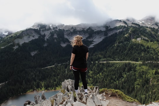 woman standing on rock cliff overlooking trees and body of water with mountains at distance during daytime in Mount Rainier National Park United States
