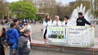 A group of young people holding a banner that pledges to restrict global warming to under 2°C. The scene takes place in an outdoor area with a fountain in the background. Several other people and camera crew are visible, indicating a public or community event focused on climate change awareness.