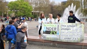 A group of young people holding a banner that pledges to restrict global warming to under 2°C. The scene takes place in an outdoor area with a fountain in the background. Several other people and camera crew are visible, indicating a public or community event focused on climate change awareness.