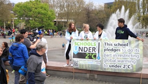 A group of young people holding a banner that pledges to restrict global warming to under 2°C. The scene takes place in an outdoor area with a fountain in the background. Several other people and camera crew are visible, indicating a public or community event focused on climate change awareness.