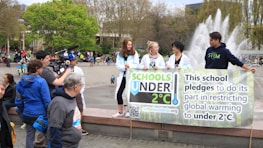 A group of young people holding a banner that pledges to restrict global warming to under 2°C. The scene takes place in an outdoor area with a fountain in the background. Several other people and camera crew are visible, indicating a public or community event focused on climate change awareness.