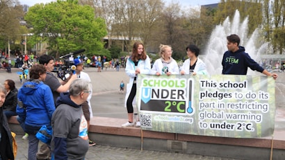 A group of young people holding a banner that pledges to restrict global warming to under 2&deg;C. The scene takes place in an outdoor area with a fountain in the background. Several other people and camera crew are visible, indicating a public or community event focused on climate change awareness.