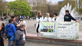 A group of young people holding a banner that pledges to restrict global warming to under 2°C. The scene takes place in an outdoor area with a fountain in the background. Several other people and camera crew are visible, indicating a public or community event focused on climate change awareness.