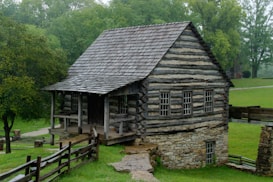 A rustic log cabin with a wooden shingle roof set in a lush green landscape. The cabin features multiple small windows and a small porch with two wooden steps leading up to it. Surrounding the cabin, a wooden fence encloses the area with trees and grass providing a serene natural backdrop.