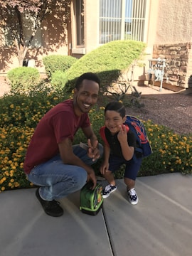 A smiling adult and child are crouching on a pavement. The adult is wearing a red shirt and jeans, showing a peace sign with one hand. The child, with a backpack, is wearing a dark outfit and sneakers, also gesturing with his hand. They are surrounded by flowering bushes in a residential area.