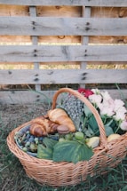 A wicker basket filled with a variety of items, including croissants, grapes, a macaron, a peach, and vibrant flowers, rests on the grass. The basket is placed against a rustic wooden pallet backdrop, adding to the natural and cozy atmosphere.