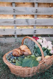 A wicker basket filled with a variety of items, including croissants, grapes, a macaron, a peach, and vibrant flowers, rests on the grass. The basket is placed against a rustic wooden pallet backdrop, adding to the natural and cozy atmosphere.
