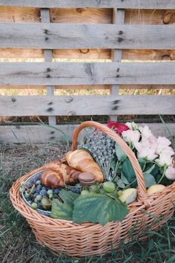 A wicker basket filled with a variety of items, including croissants, grapes, a macaron, a peach, and vibrant flowers, rests on the grass. The basket is placed against a rustic wooden pallet backdrop, adding to the natural and cozy atmosphere.