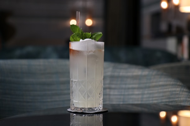 A tall, transparent glass filled with a light-colored cocktail, garnished with fresh mint leaves. The drink is served on a black coaster and has a straw. In the background, there is a blurred setting with soft, warm lighting and plush seating.
