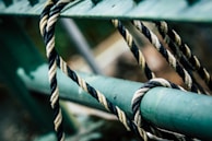 A close-up of a twisted rope looped around and loosely tied to a green metal bar. The rope features intertwined black and white strands, creating a pattern of contrast against the smooth, painted metal surface.