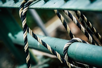 A close-up of a twisted rope looped around and loosely tied to a green metal bar. The rope features intertwined black and white strands, creating a pattern of contrast against the smooth, painted metal surface.