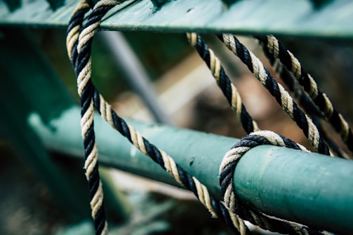 A close-up of a twisted rope looped around and loosely tied to a green metal bar. The rope features intertwined black and white strands, creating a pattern of contrast against the smooth, painted metal surface.