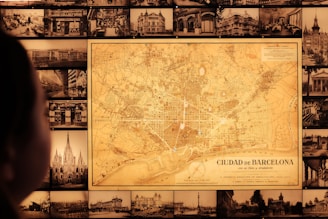 A vintage map of Barcelona is prominently displayed on a wall surrounded by numerous black-and-white historical photographs of cityscapes and buildings. A person is visible in the foreground, gazing at the map.