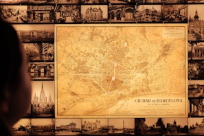 A vintage map of Barcelona is prominently displayed on a wall surrounded by numerous black-and-white historical photographs of cityscapes and buildings. A person is visible in the foreground, gazing at the map.