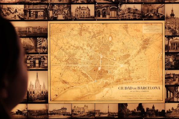 A vintage map of Barcelona is prominently displayed on a wall surrounded by numerous black-and-white historical photographs of cityscapes and buildings. A person is visible in the foreground, gazing at the map.