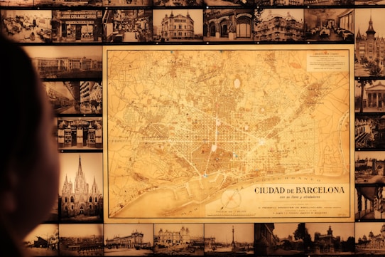 A vintage map of Barcelona is prominently displayed on a wall surrounded by numerous black-and-white historical photographs of cityscapes and buildings. A person is visible in the foreground, gazing at the map.