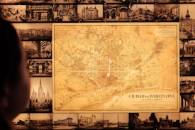 A vintage map of Barcelona is prominently displayed on a wall surrounded by numerous black-and-white historical photographs of cityscapes and buildings. A person is visible in the foreground, gazing at the map.