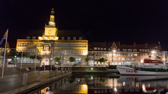 A waterfront scene at night featuring a large, illuminated building with a clock tower, reflecting in the calm water. The building is surrounded by smaller structures with a row of streetlamps casting a warm glow. A rescue boat labeled 'SAR II' is docked along the edge of the water, with flags waving on poles nearby.