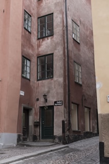 A narrow cobblestone street leads to a corner building with a rustic appearance. The building features weathered brown and pink walls and several windows with dark frames. A green door marked 'Galleri Norrsken' is prominently visible. The street and building convey a historic and European architectural style.