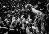 A large group of people is gathered in a lively and celebratory atmosphere. Confetti is raining down from above, and many individuals are smiling and holding up their phones, capturing the moment. The lighting is dynamic, adding to the energetic vibe of the scene.