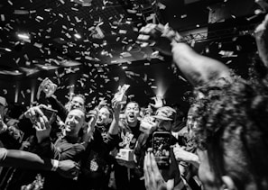A large group of people is gathered in a lively and celebratory atmosphere. Confetti is raining down from above, and many individuals are smiling and holding up their phones, capturing the moment. The lighting is dynamic, adding to the energetic vibe of the scene.