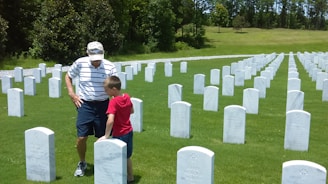 An elderly man and a young boy stand among rows of white headstones in a neatly maintained cemetery. They appear to be observing one of the gravestones together. The surroundings are lush with green grass and a backdrop of trees.