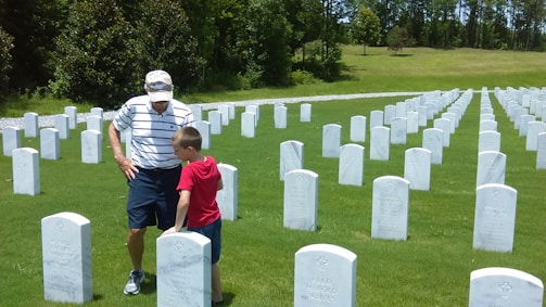 An elderly man and a young boy stand among rows of white headstones in a neatly maintained cemetery. They appear to be observing one of the gravestones together. The surroundings are lush with green grass and a backdrop of trees.