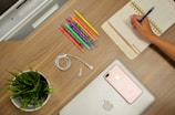 A neatly organized wooden desk features a person writing in a planner with a pen, surrounded by colorful pens, a potted green plant, a coiled pair of white earphones, a smartphone, and a closed laptop with an Apple logo.