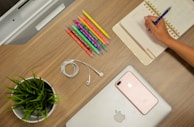 A neatly organized wooden desk features a person writing in a planner with a pen, surrounded by colorful pens, a potted green plant, a coiled pair of white earphones, a smartphone, and a closed laptop with an Apple logo.