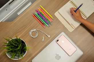 A neatly organized wooden desk features a person writing in a planner with a pen, surrounded by colorful pens, a potted green plant, a coiled pair of white earphones, a smartphone, and a closed laptop with an Apple logo.