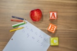 A red apple lies next to colorful alphabet blocks labeled A, B, and C on a wooden surface. Nearby, there is a grid paper with handwritten letters of the alphabet in blue ink and several colored pencils in shades of green, blue, red, white, and yellow.