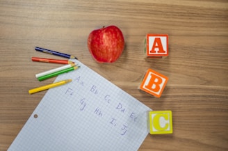 A red apple lies next to colorful alphabet blocks labeled A, B, and C on a wooden surface. Nearby, there is a grid paper with handwritten letters of the alphabet in blue ink and several colored pencils in shades of green, blue, red, white, and yellow.