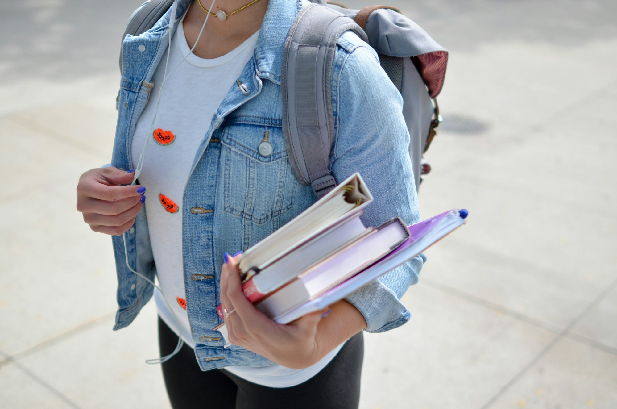 Fille habillée avec une veste en jean et un T-shirt blanc et portant une pile de livre dans le bras et un sac à dos gris