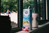 A carton of Dutch Lady PureFarm milk and a can of another beverage rest on a windowsill. The carton has a straw inserted, and there is an empty candy wrapper in front. Outside the window, blurred figures of people and greenery are visible, creating a casual, everyday scene.