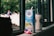 A carton of Dutch Lady PureFarm milk and a can of another beverage rest on a windowsill. The carton has a straw inserted, and there is an empty candy wrapper in front. Outside the window, blurred figures of people and greenery are visible, creating a casual, everyday scene.