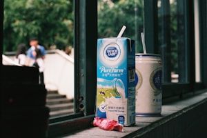A carton of Dutch Lady PureFarm milk and a can of another beverage rest on a windowsill. The carton has a straw inserted, and there is an empty candy wrapper in front. Outside the window, blurred figures of people and greenery are visible, creating a casual, everyday scene.