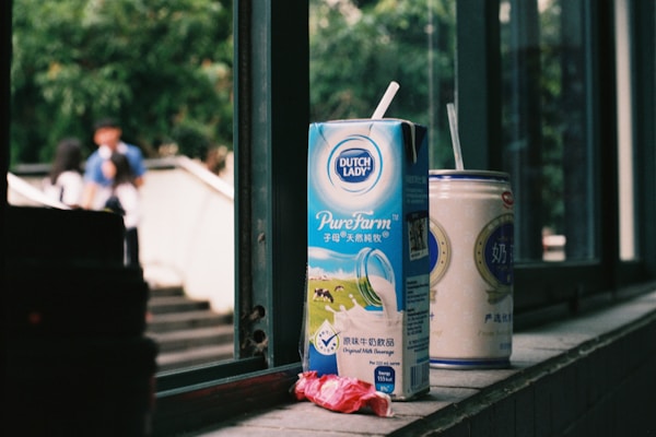 A carton of Dutch Lady PureFarm milk and a can of another beverage rest on a windowsill. The carton has a straw inserted, and there is an empty candy wrapper in front. Outside the window, blurred figures of people and greenery are visible, creating a casual, everyday scene.