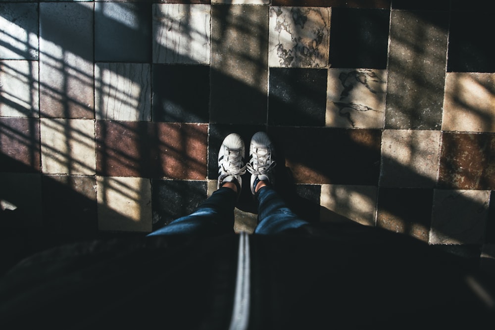 high-angle of person in blue pants and white sneakers standing on floor tiles