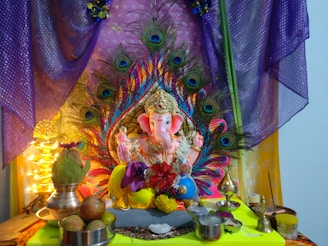 A colorful and detailed Ganesha idol is surrounded by vibrant peacock feathers. The setup includes various offerings like fruits in bowls and a coconut placed next to the idol. The background is adorned with purple and gold fabric, and small lights add a warm glow. The scene is rich in traditional and cultural elements.