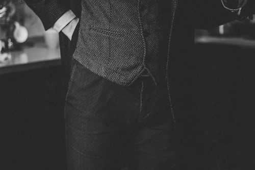A black and white image showing a person dressed in a textured suit with their hand in their pocket. The focus is on the midsection of the person, highlighting the details of the suit fabric and the casual yet sophisticated posture.