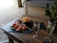 A wooden table set with two glasses of sparkling wine and a plate of bruschetta topped with chopped tomatoes and greens. Next to it is a dish with cheese cubes. There's a small plant in a woven basket, and a cushioned seat in the background.