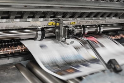 A high-speed printing press processes sheets of paper rapidly, with machinery featuring spinning rollers and metallic components. Blurred motion suggests continuous production, and a caution label is visible on the equipment.