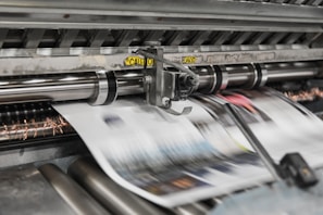 A high-speed printing press processes sheets of paper rapidly, with machinery featuring spinning rollers and metallic components. Blurred motion suggests continuous production, and a caution label is visible on the equipment.