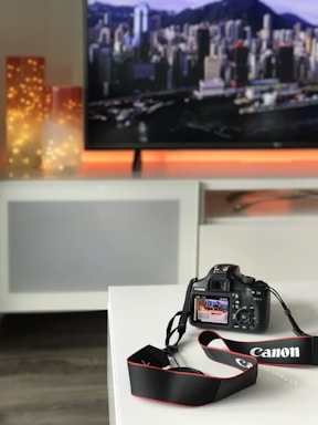 A digital camera with a Canon logo sits on a white surface. In the background, there are illuminated decorative lights and a large screen displaying a blurred cityscape. The camera's display shows part of the scene, adding depth to the composition.
