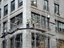 Three construction workers are on scaffolding attached to a tall building. They are wearing high-visibility vests and hard hats while working on renovations or maintenance of the structure. The building appears to be made of stone or concrete with large rectangular windows.
