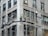 Three construction workers are on scaffolding attached to a tall building. They are wearing high-visibility vests and hard hats while working on renovations or maintenance of the structure. The building appears to be made of stone or concrete with large rectangular windows.