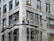 Three construction workers are on scaffolding attached to a tall building. They are wearing high-visibility vests and hard hats while working on renovations or maintenance of the structure. The building appears to be made of stone or concrete with large rectangular windows.
