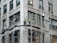 Three construction workers are on scaffolding attached to a tall building. They are wearing high-visibility vests and hard hats while working on renovations or maintenance of the structure. The building appears to be made of stone or concrete with large rectangular windows.