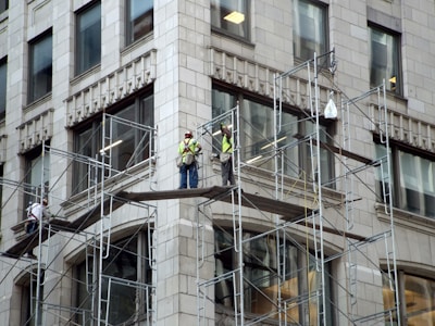 Three construction workers are on scaffolding attached to a tall building. They are wearing high-visibility vests and hard hats while working on renovations or maintenance of the structure. The building appears to be made of stone or concrete with large rectangular windows.