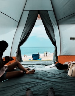 Inside view of a tent with the entrance open, revealing a sandy beach in the background. The ocean is visible under a clear sky. A person is sitting inside the tent with a camera, and there are several items placed around such as a bag and some clothes. People are visible walking near the water and a blue beach chair is set up at the shoreline.